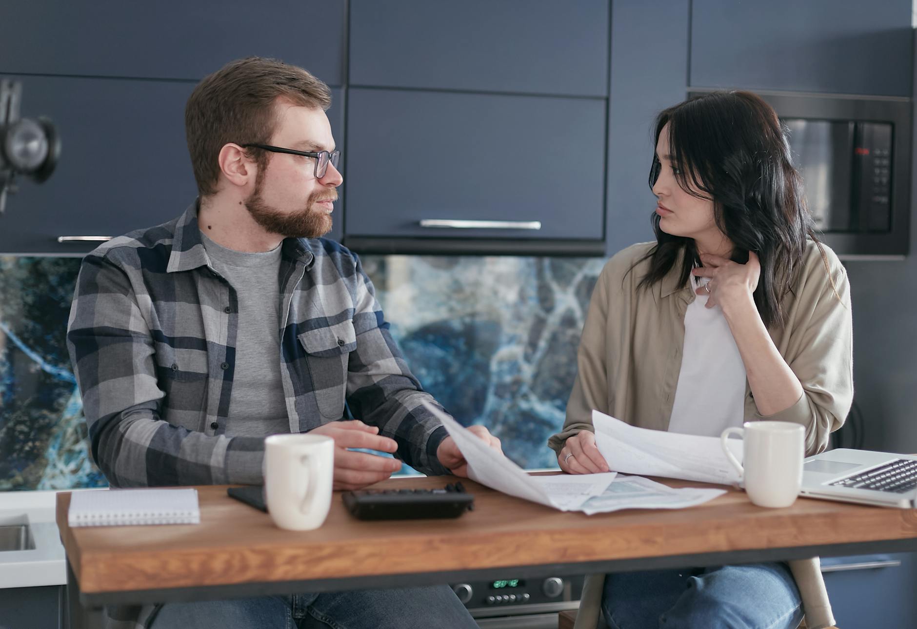 A young couple sitting at a table discussing bills and financial plans in a modern kitchen. - couples financial stress communication