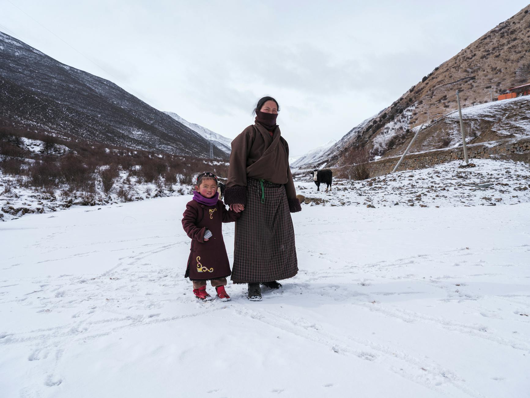 A mother and daughter in traditional clothing stand on a snowy landscape in a rural winter setting. - family snow day communication