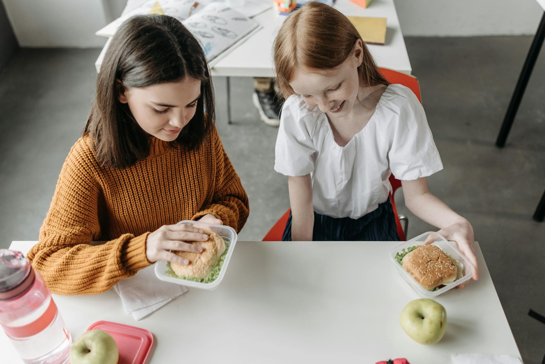 Two young girls enjoying lunch together at a school desk with sandwiches and apples. - february school break parenting