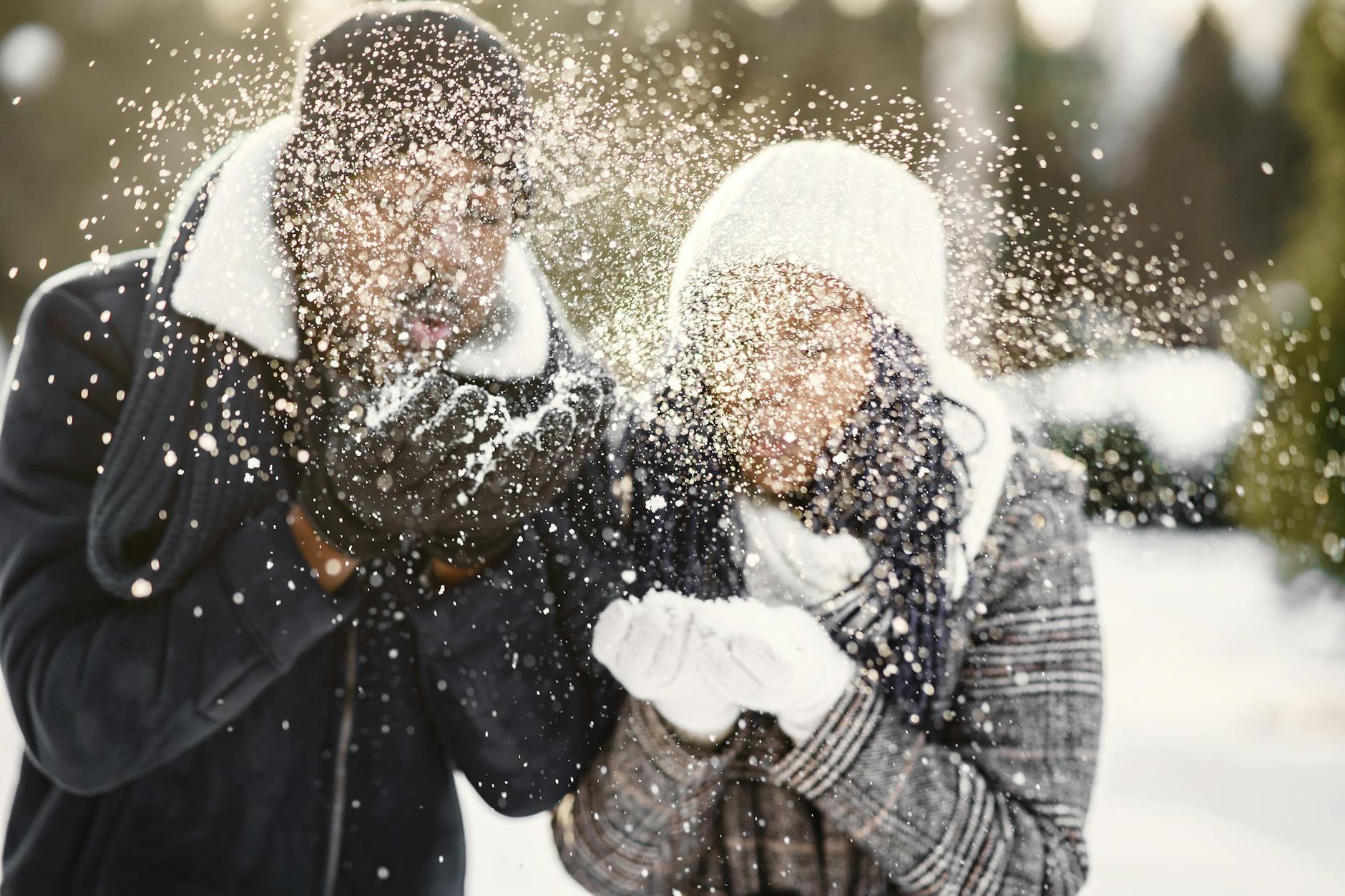 A joyful couple blowing snowflakes with winter apparel, embodying playful winter bliss. - intimate winter date ideas