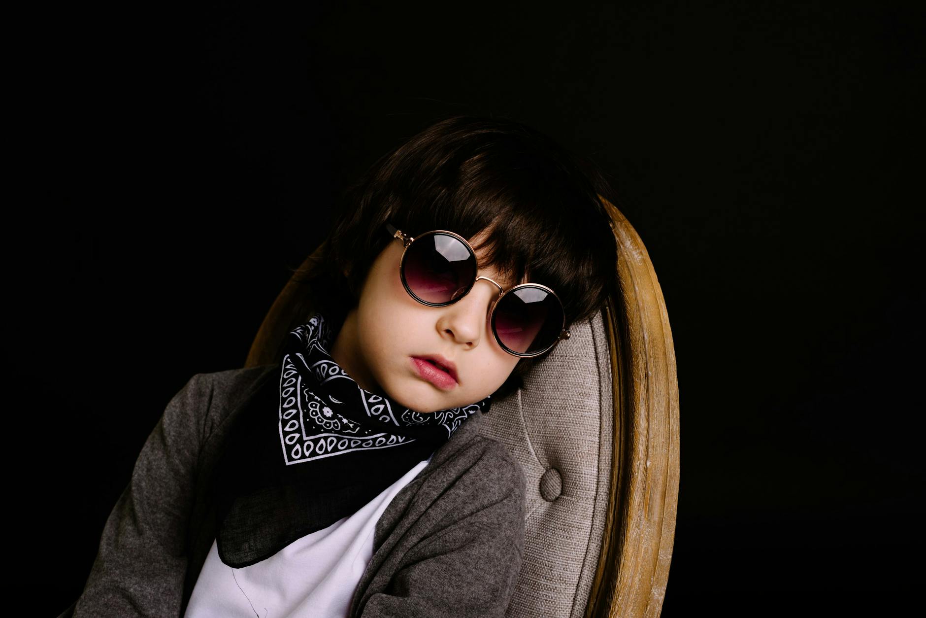 Portrait of a fashionable child wearing sunglasses and a scarf, sitting on a chair against a dark background. - kids winter break boredom
