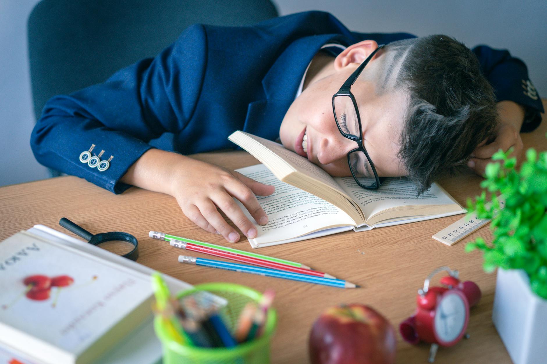 A tired schoolboy in glasses sleeping on a desk surrounded by study materials. - kids winter break boredom