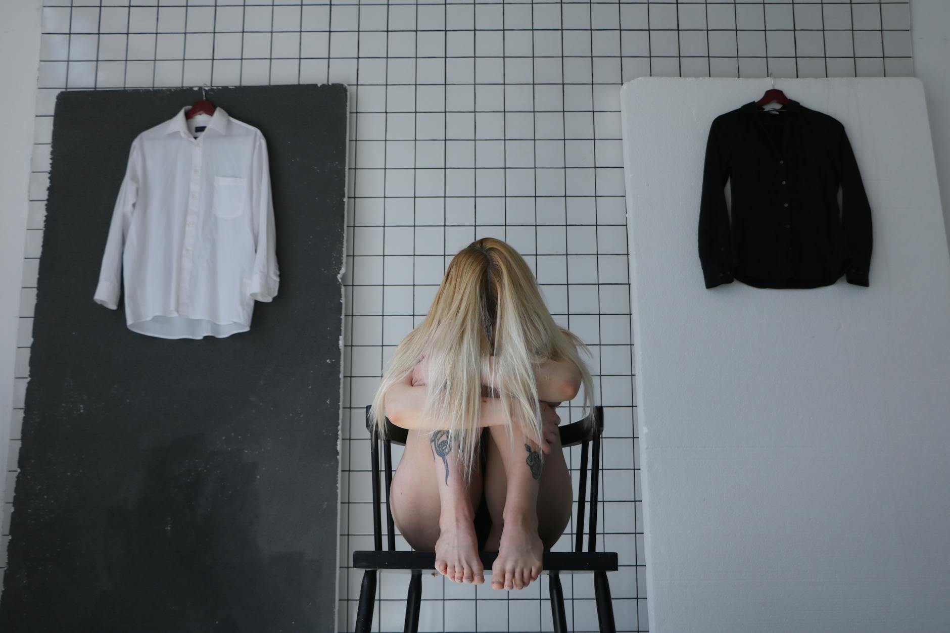 Unrecognizable barefoot depressed tattooed female embracing knees while sitting on stool behind black and white shirts on hangers - mindfulness for winter anxiety
