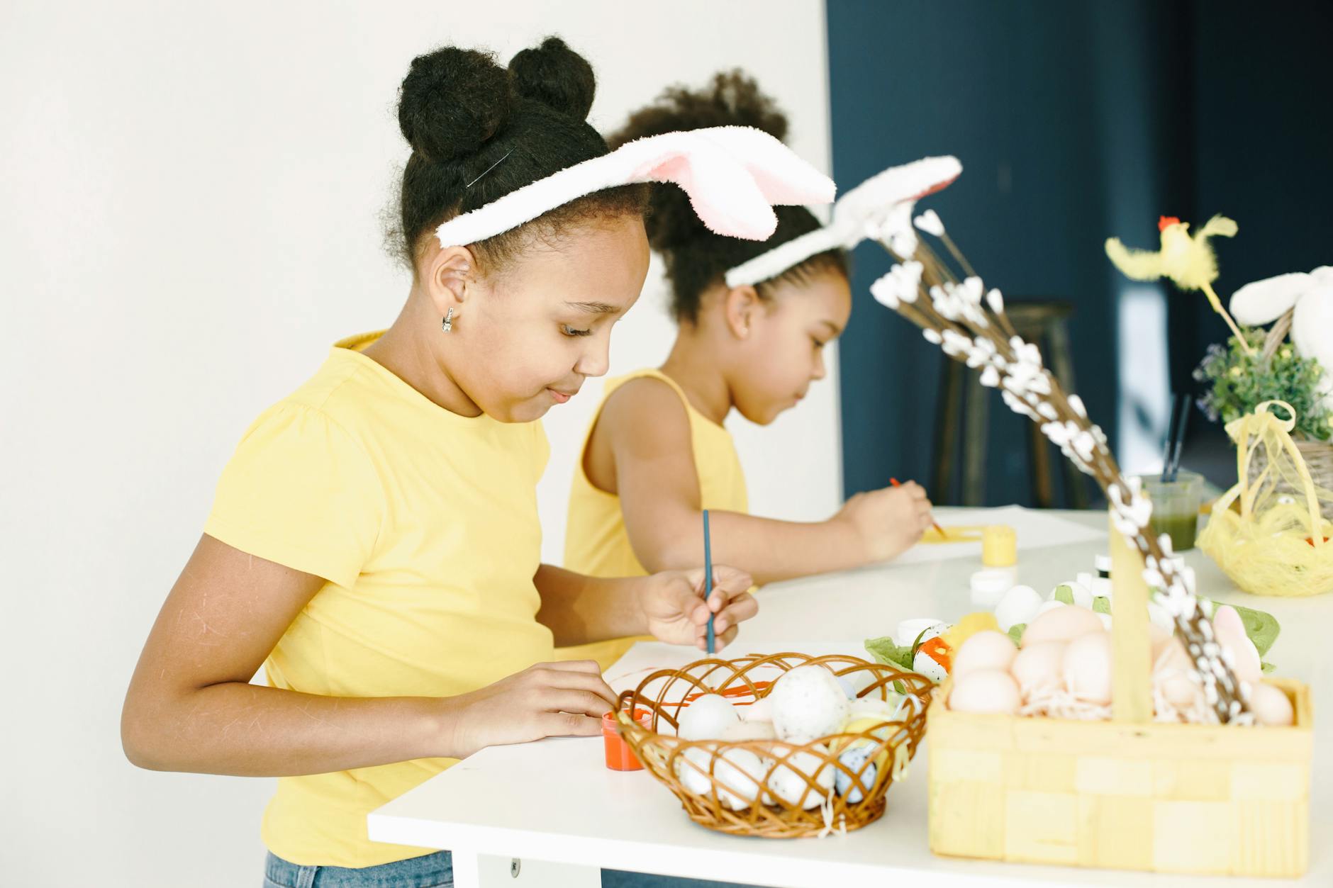 Girls painting Easter eggs indoors, wearing bunny ears and yellow outfits, having fun. - preparing kids for spring transition