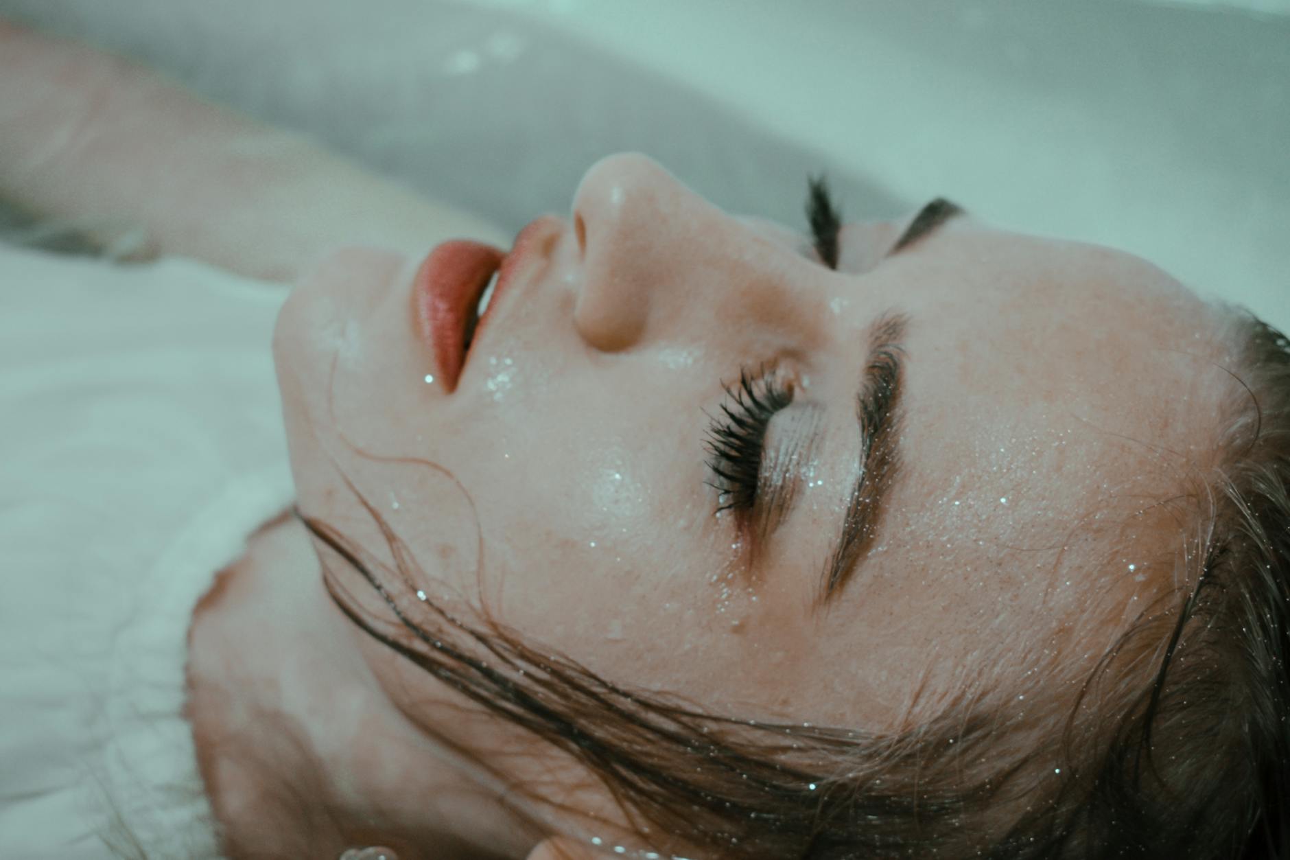 Relaxed female wearing white outfit with wet hair lying in bathtub in water with closed eyes at home in bathroom - seasonal depression teens
