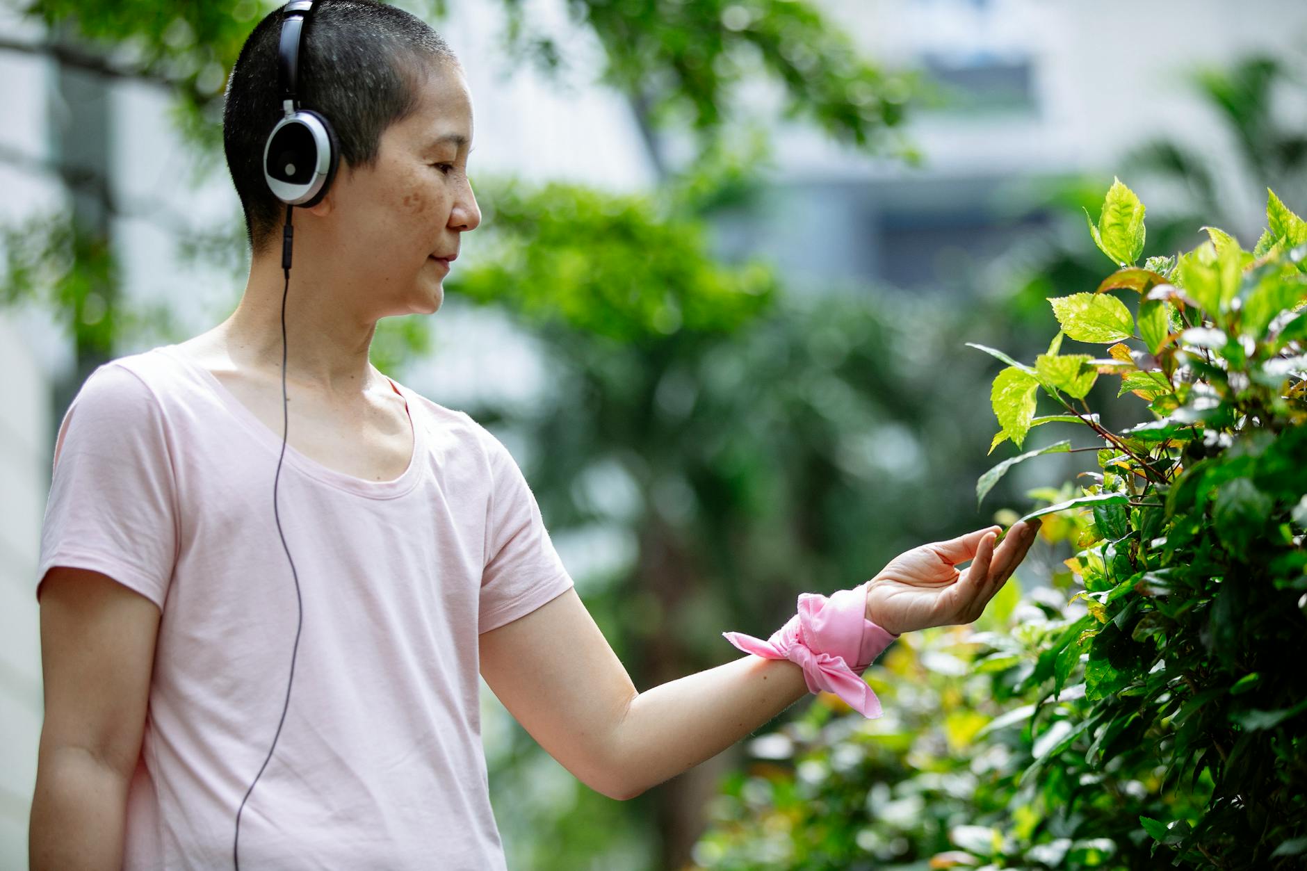 Thoughtful middle age ethnic woman in casual clothes with headphones and pink bracelet examining bushes and plants with green leaves in park in summer day - active listening examples