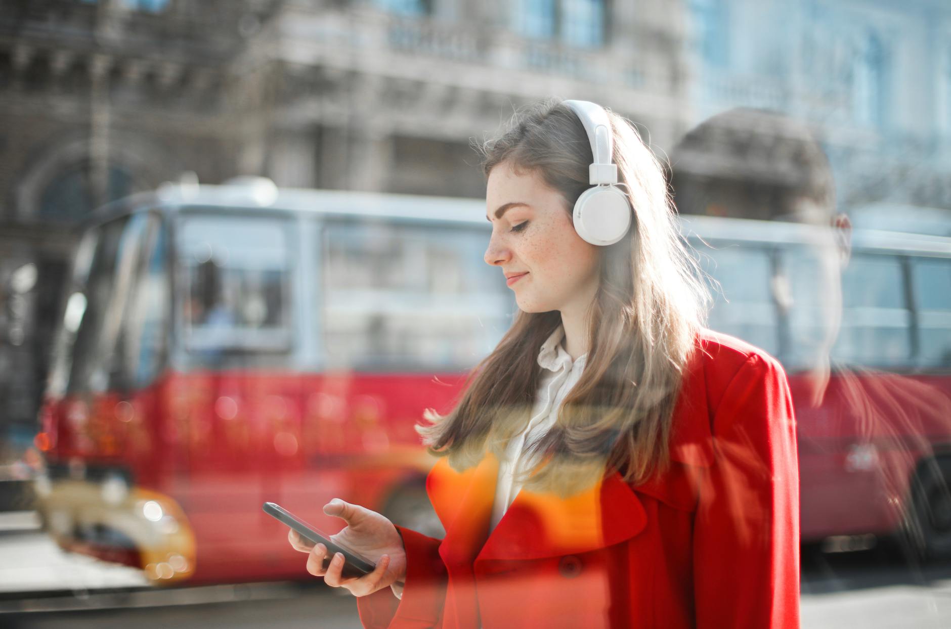Young woman in red blazer using smartphone and headphones outdoors with bus in background. - active listening examples
