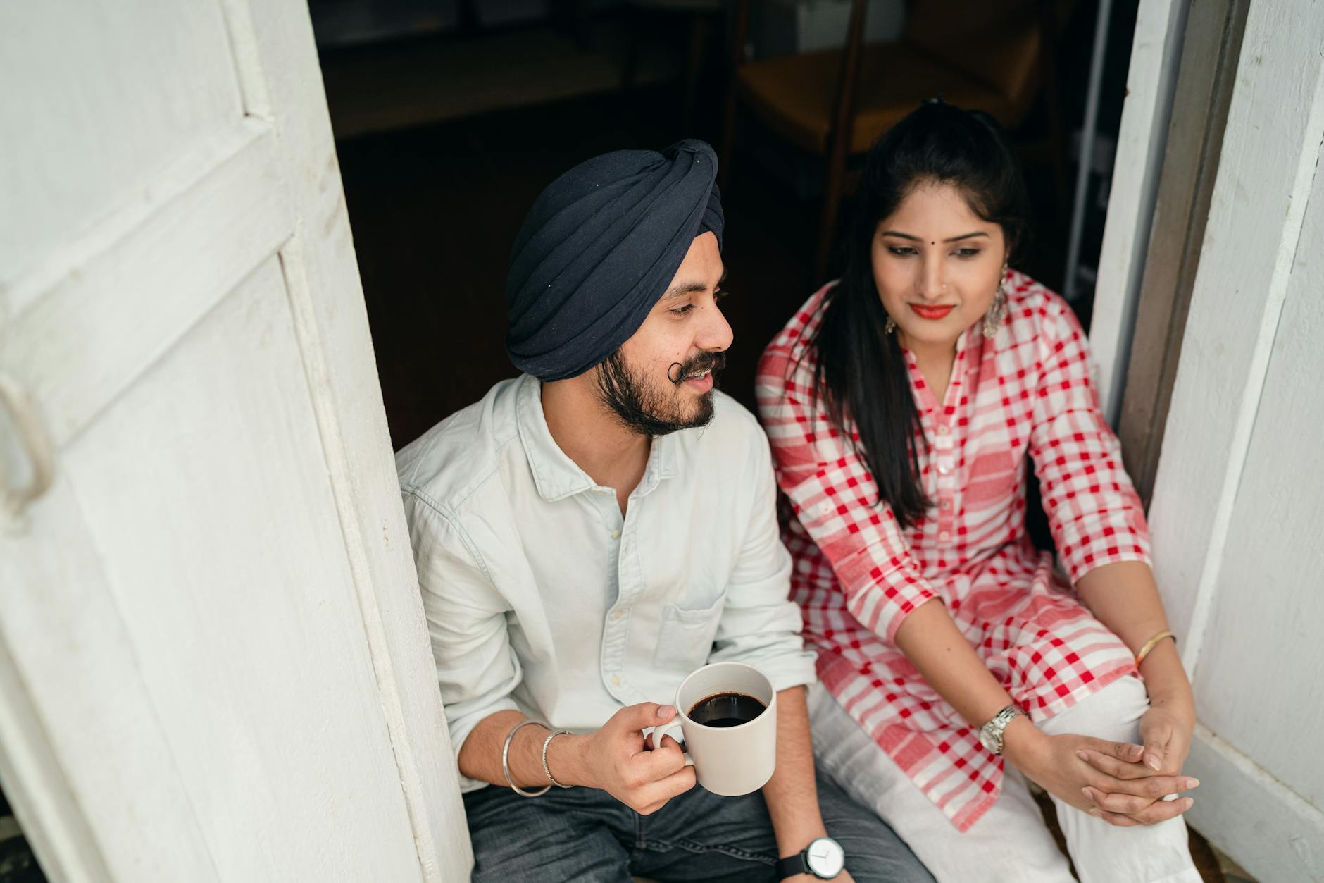 From above of good looking Indian wife listening to husband sharing news with cup of coffee in hand while both sitting at doorstep of house - active listening relationship
