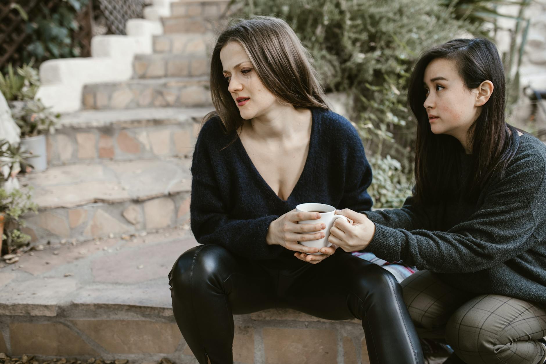 Two women share emotional support on outdoor steps, one consoling the other with a cup. - active listening steps