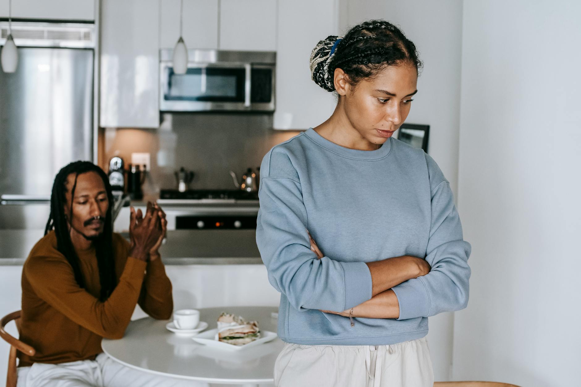 Young discontented African American female with folded arms against male partner at kitchen table during quarrel - allergy irritability relationship