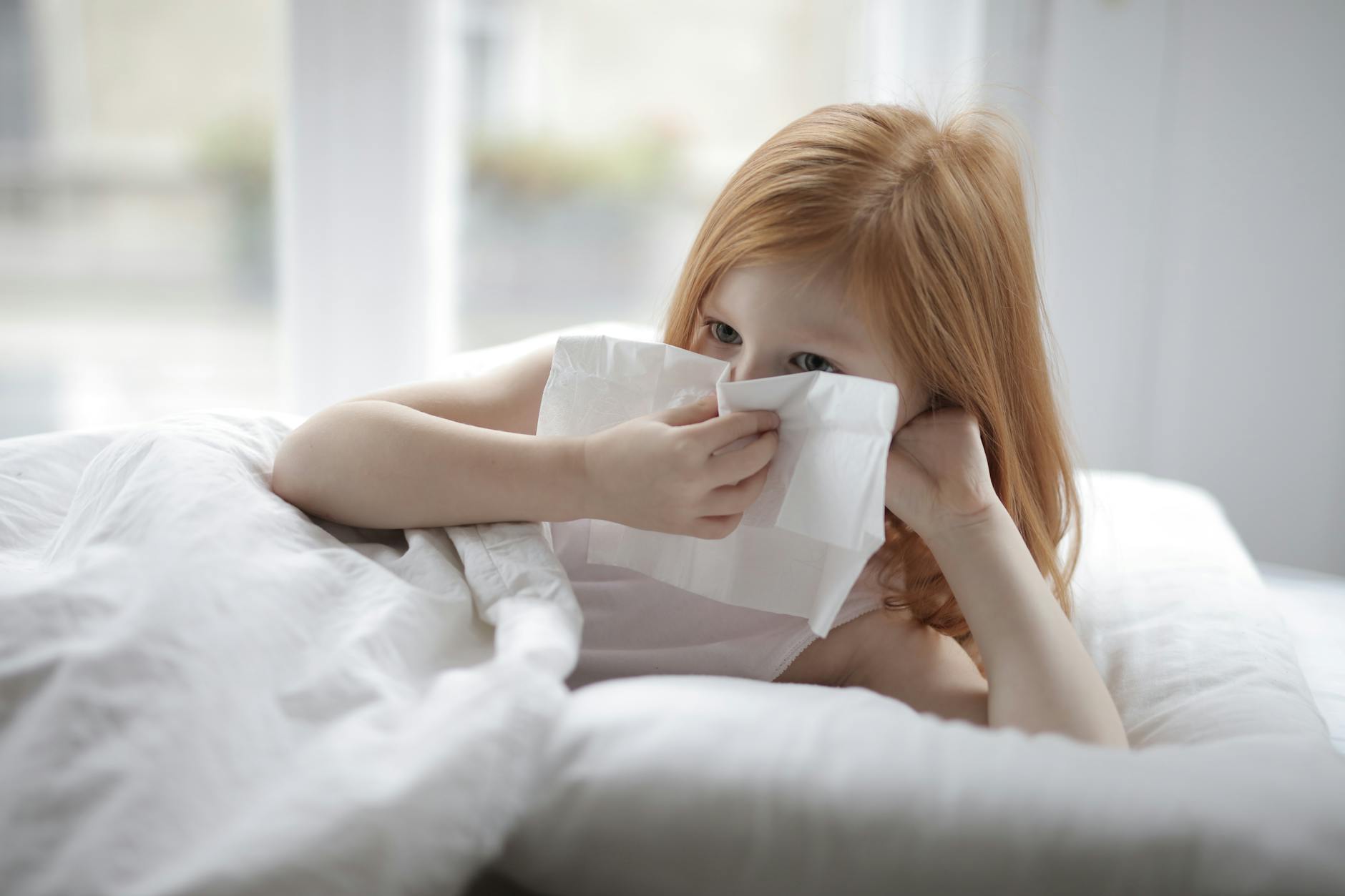 Cute unhealthy child with red hair blowing nose with paper towel while lying on bed with white linen in cozy light bedroom - allergy mood changes