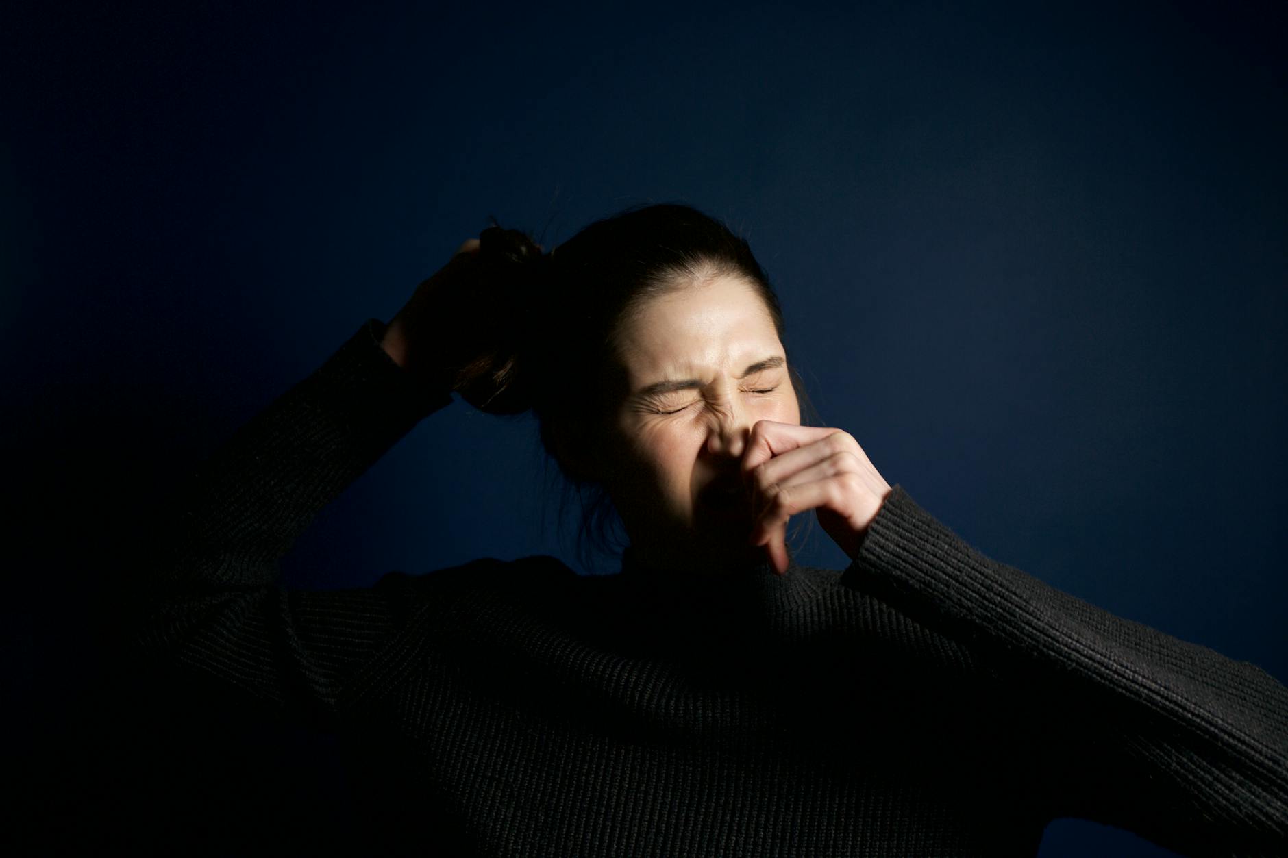 A woman in a dark sweater sneezes against a solid blue background, capturing a moment of candid expression. - allergy relationship strain
