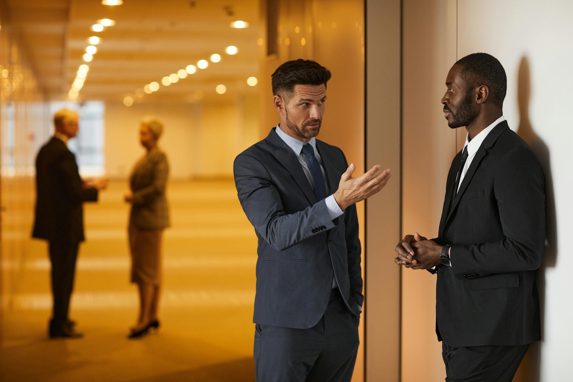 Two businessmen in formal attire engaged in a serious discussion in a modern office hallway. - assertive communication meaning