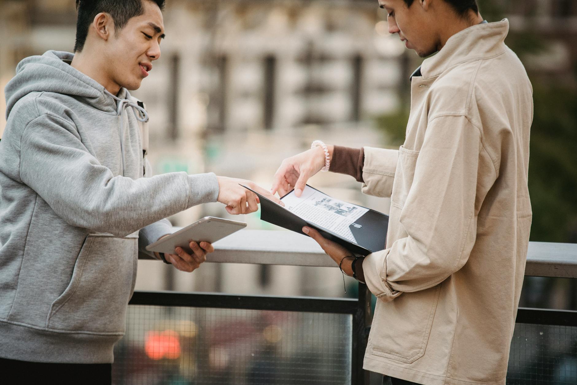 Side view of young diverse male students in casual clothes standing on street with tablet and papers in folder and speaking about home task - assertive communication skills