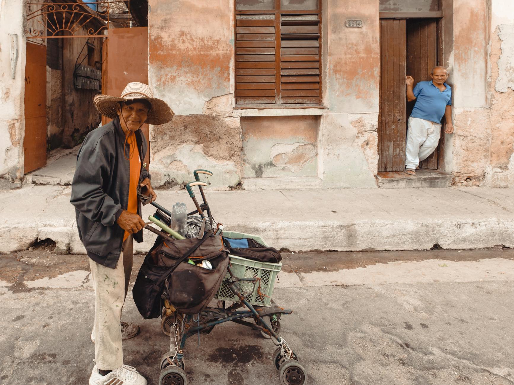 An elderly street vendor with a hat smiles while pushing a cart along a weathered building, depicting urban life. - building resilience