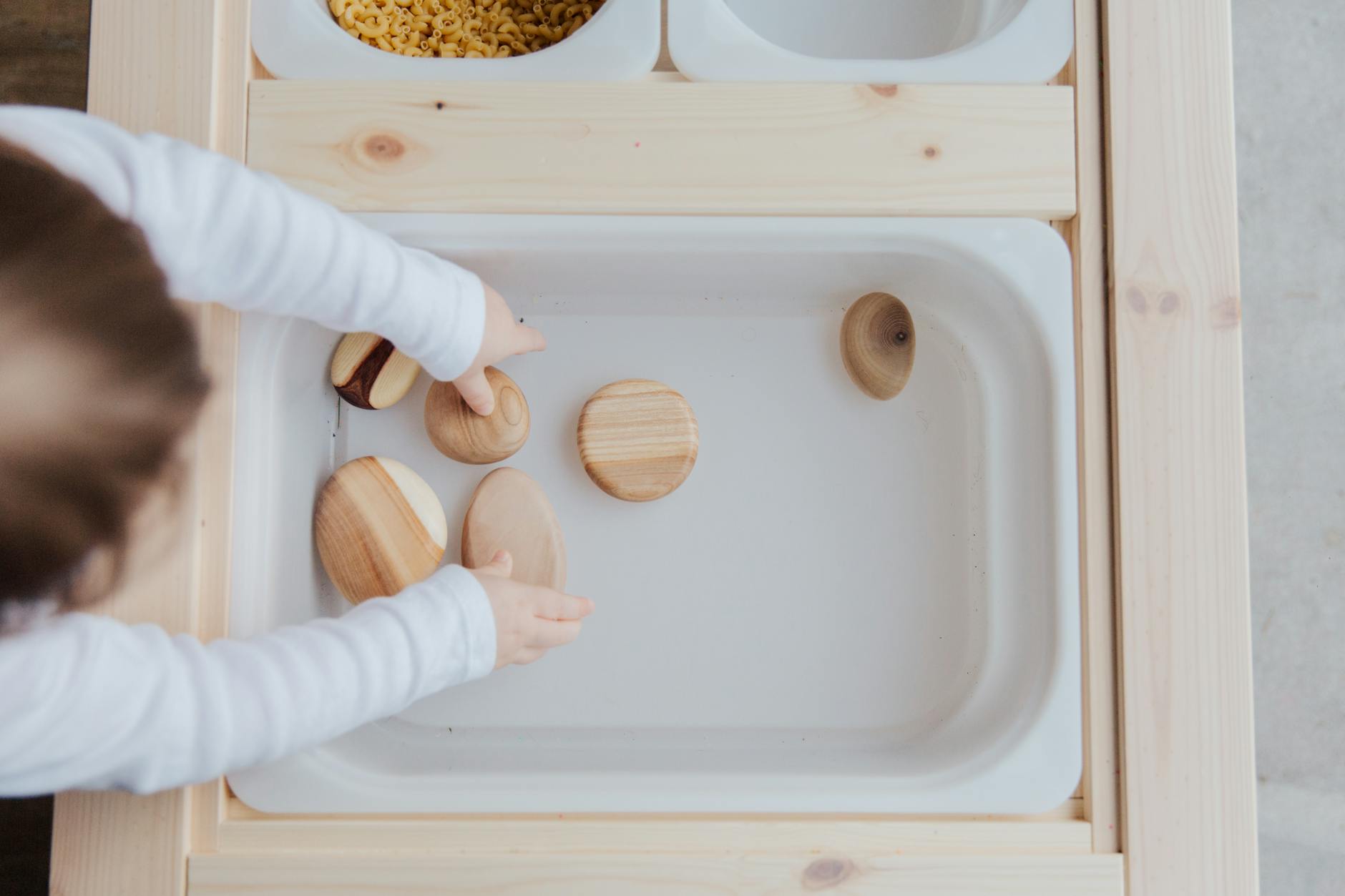 From above view of unrecognizable little child playing with wooden round stones in white plastic container at wooden table at home - child developmental milestones