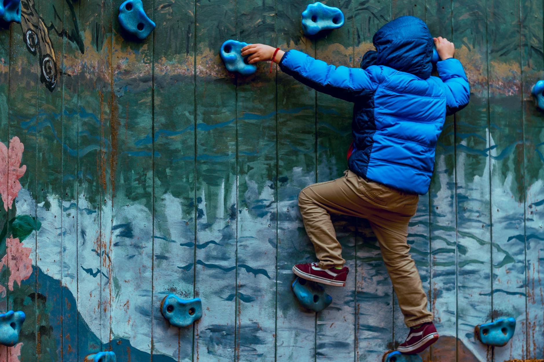 Young boy in blue jacket climbing a painted rock wall outdoors, showcasing adventure and play. - child growth spurts