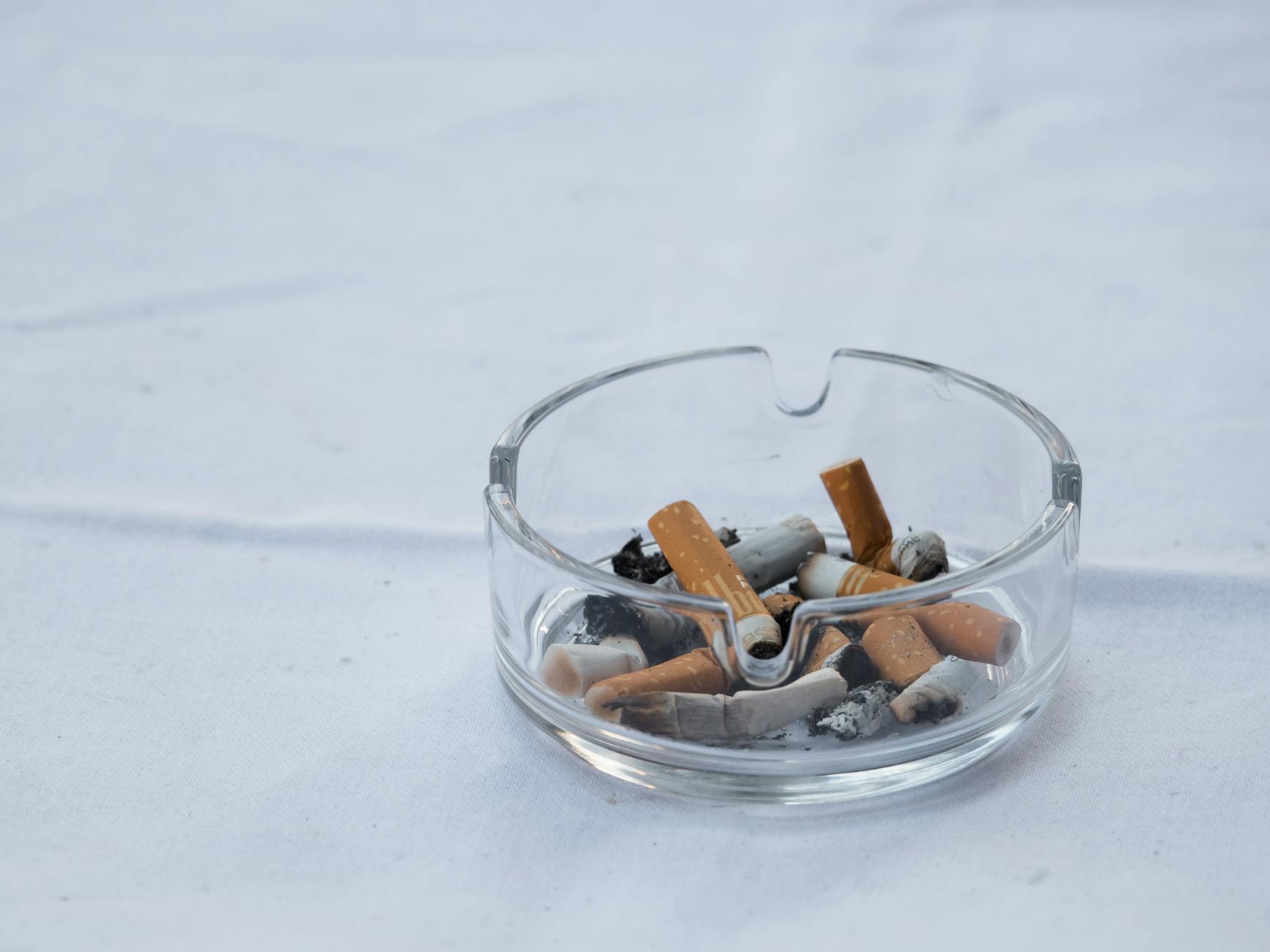 A clear glass ashtray filled with cigarette butts and ash placed on a white surface. - communication habits