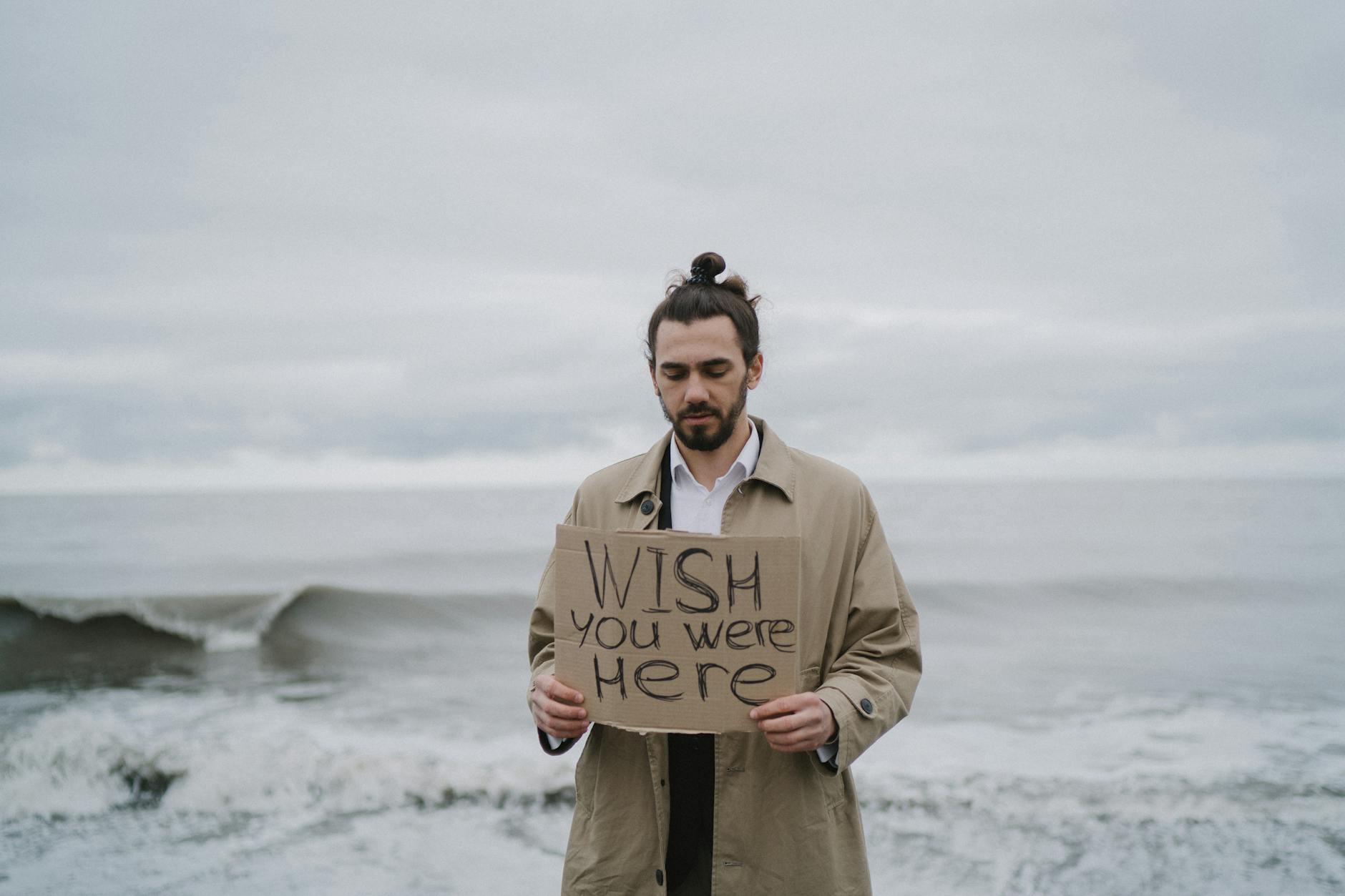 A man in a trench coat stands by the sea holding a 'Wish you were here' sign, expressing longing. - couple resentment spring