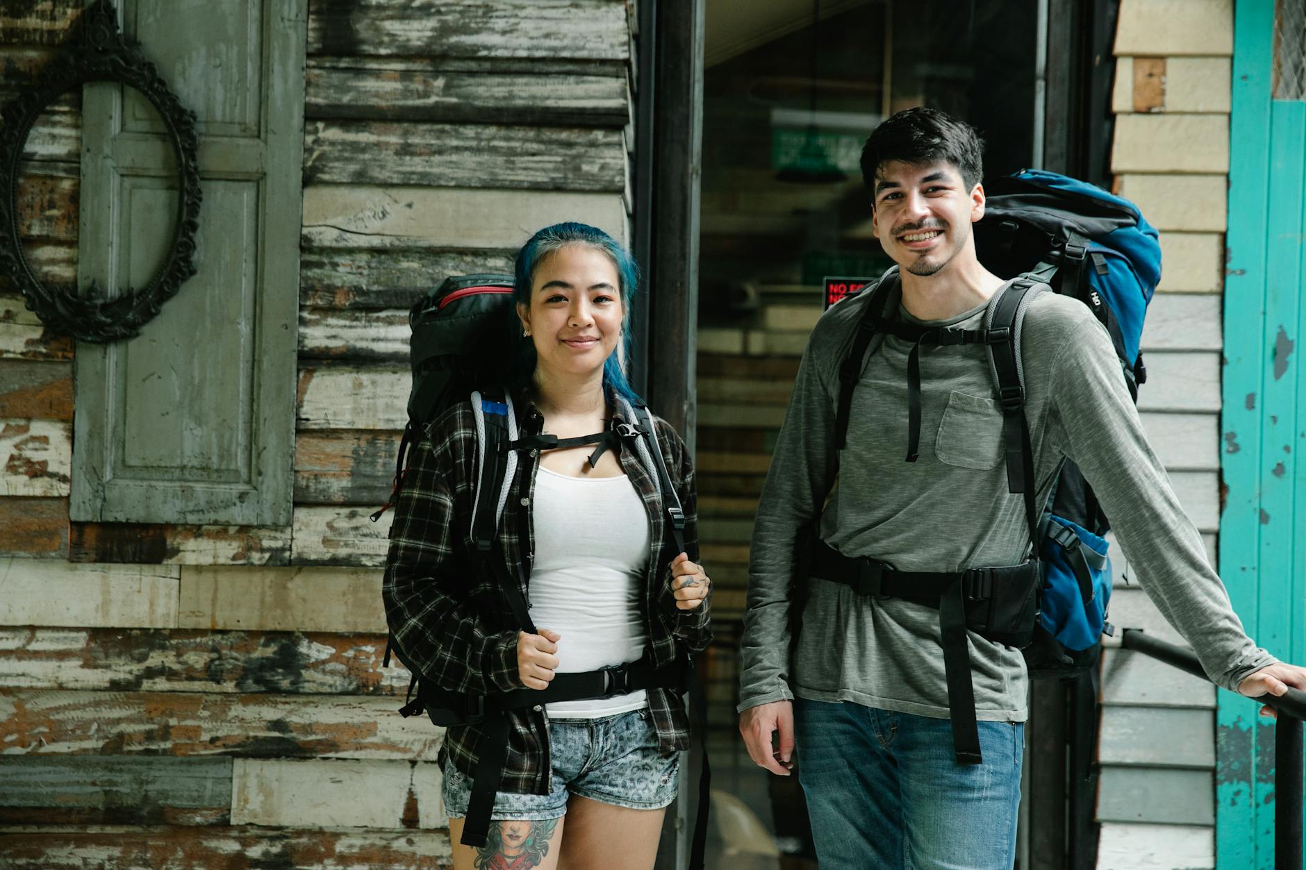Young smiling diverse couple of travelers in casual clothes and with backpacks standing together near wooden wall in light room and looking at camera - couple travel planning communication