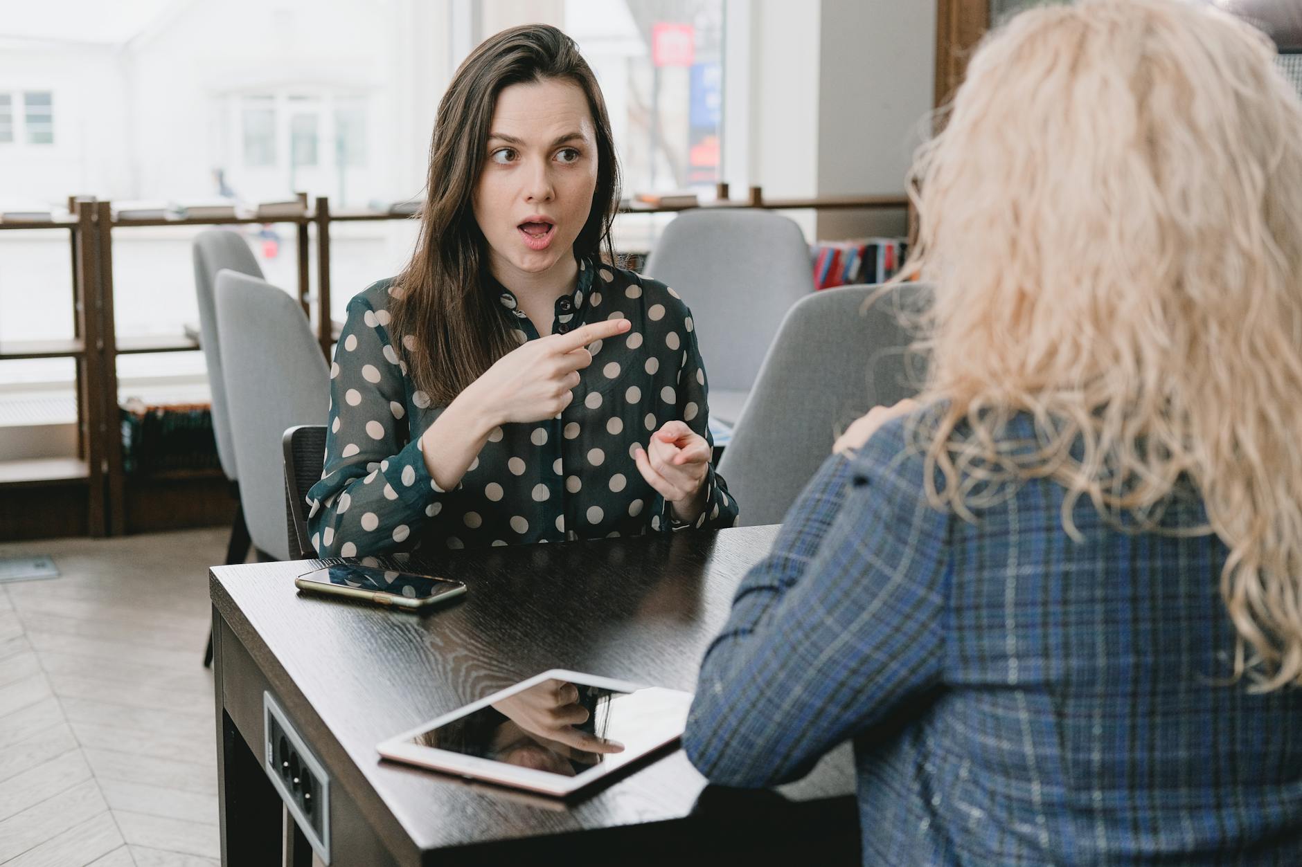 Two women in a lively discussion at a café, with expressive hand gestures and mobile devices on the table. - define assertive communication