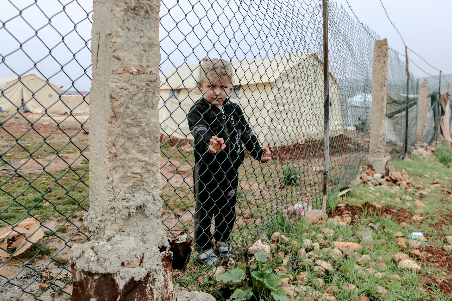 A young child stands behind a fence with tents visible in a Syrian refugee camp, conveying a poignant moment. - emotional resilience children