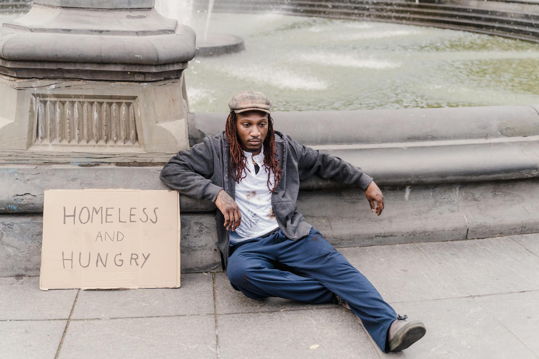 A man sits by a fountain with a cardboard sign reading 'homeless and hungry'. - express needs clearly