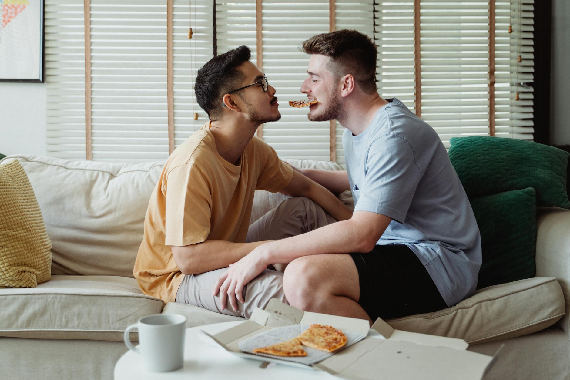 Interracial gay couple sharing pizza on sofa, expressing love and togetherness. - express needs couples