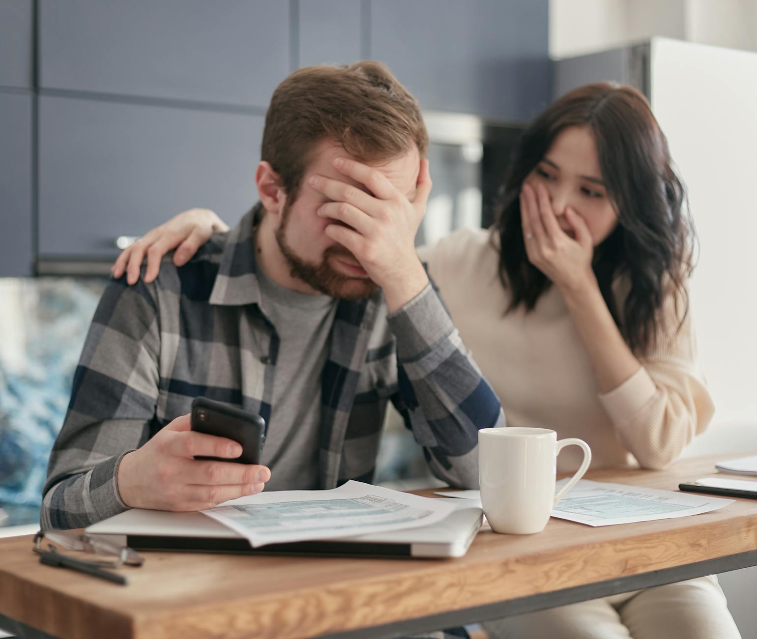 A couple looking stressed and holding a cellphone while reviewing financial documents indoors. Hands on faces in concern. - financial stress couple