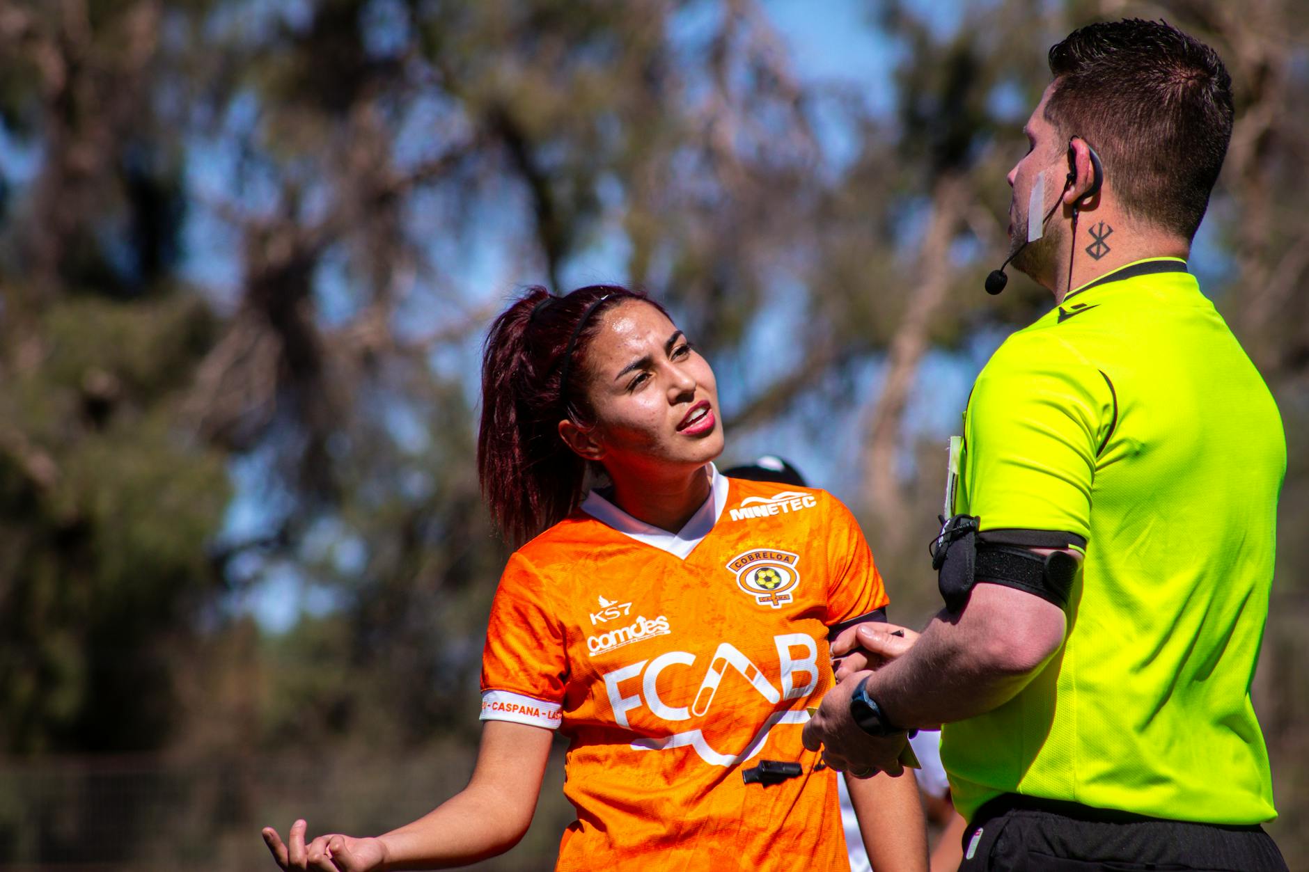 Female soccer player in orange jersey argues with referee on the outdoor field during a sunny day match. - future goals partner