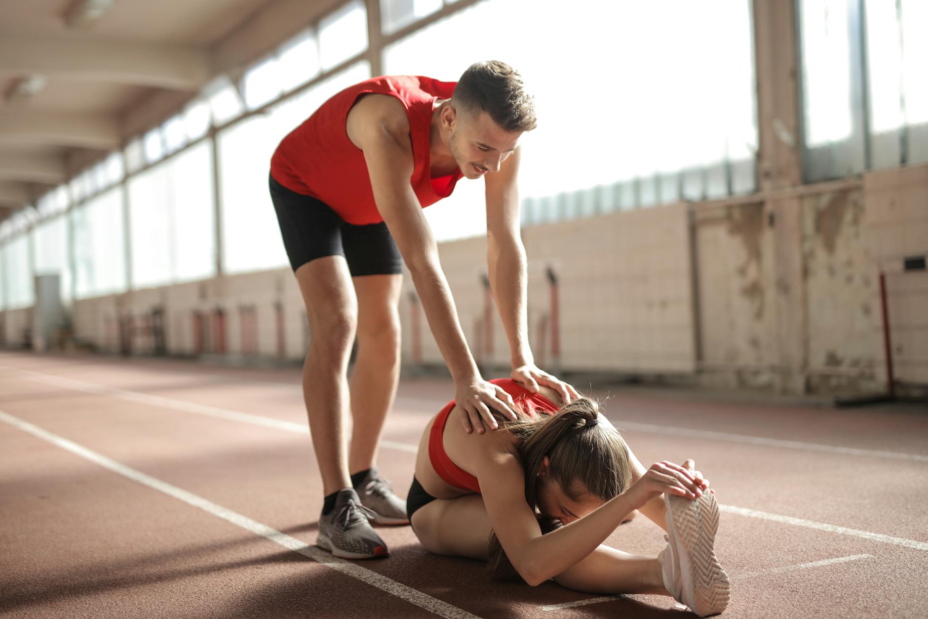 Professional male sportsman helping woman touching knees with head while female sitting on running track and doing stretching exercise - future goals partner