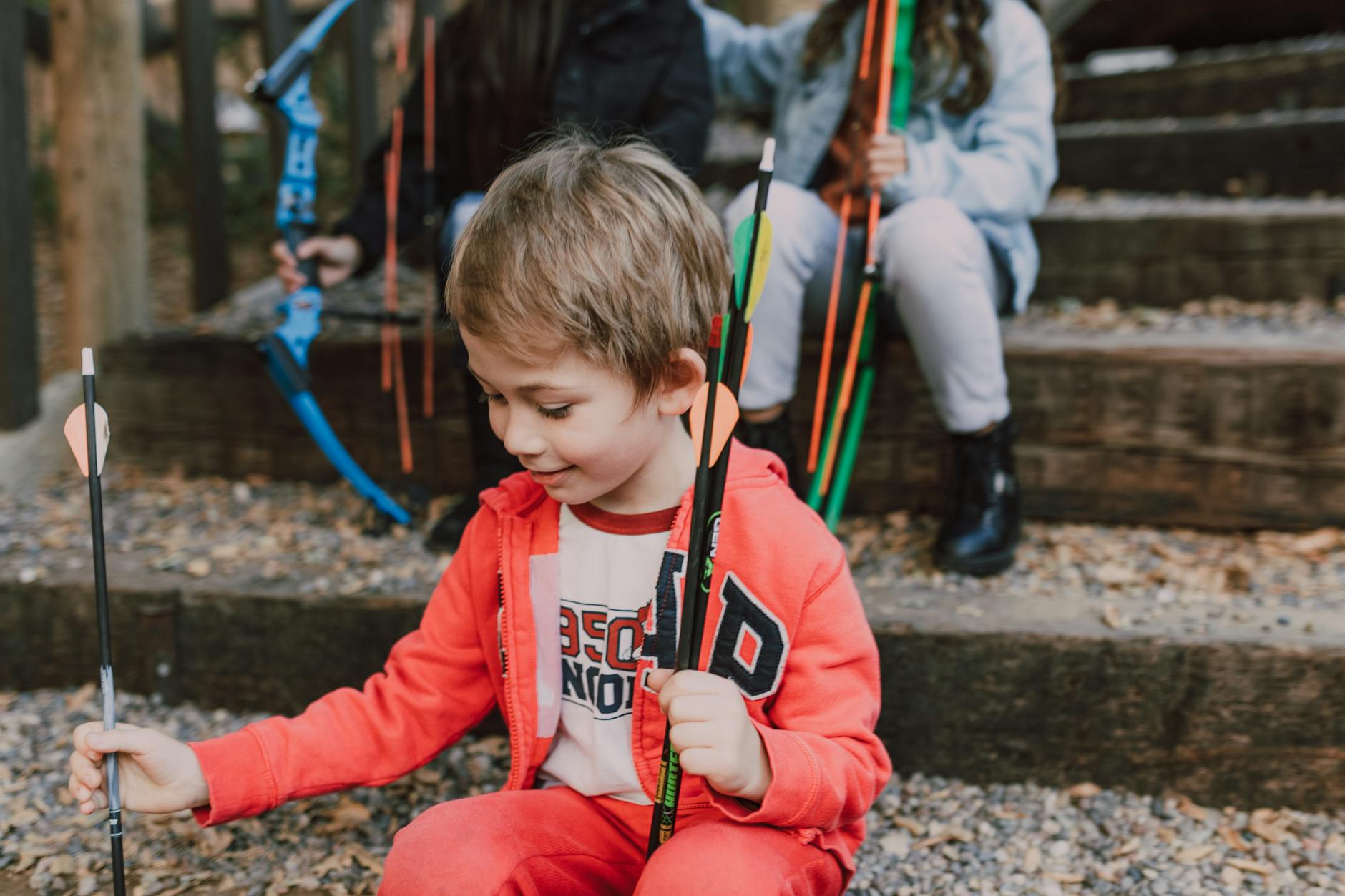 A young boy in a red outfit holds arrows happily while sitting outdoors. Engaging archery activity. - kids nature exploration