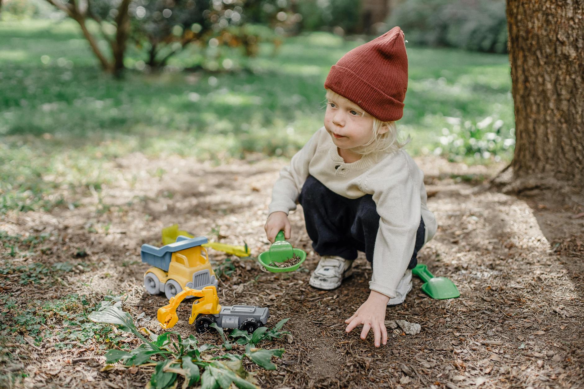 A child wearing a red beanie plays with toy trucks outdoors on a sunny day in a park. - kids nature exploration