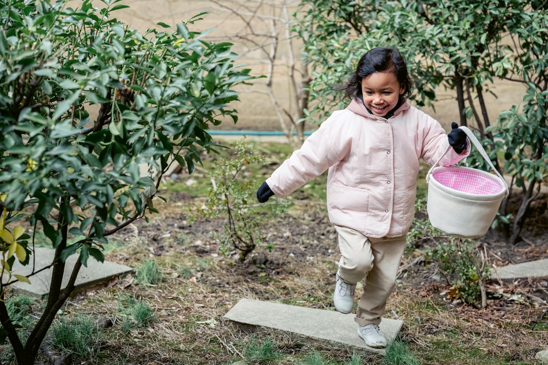 A cheerful child in a pink coat enjoying a playful day outdoors with a basket. - kids spring gardening