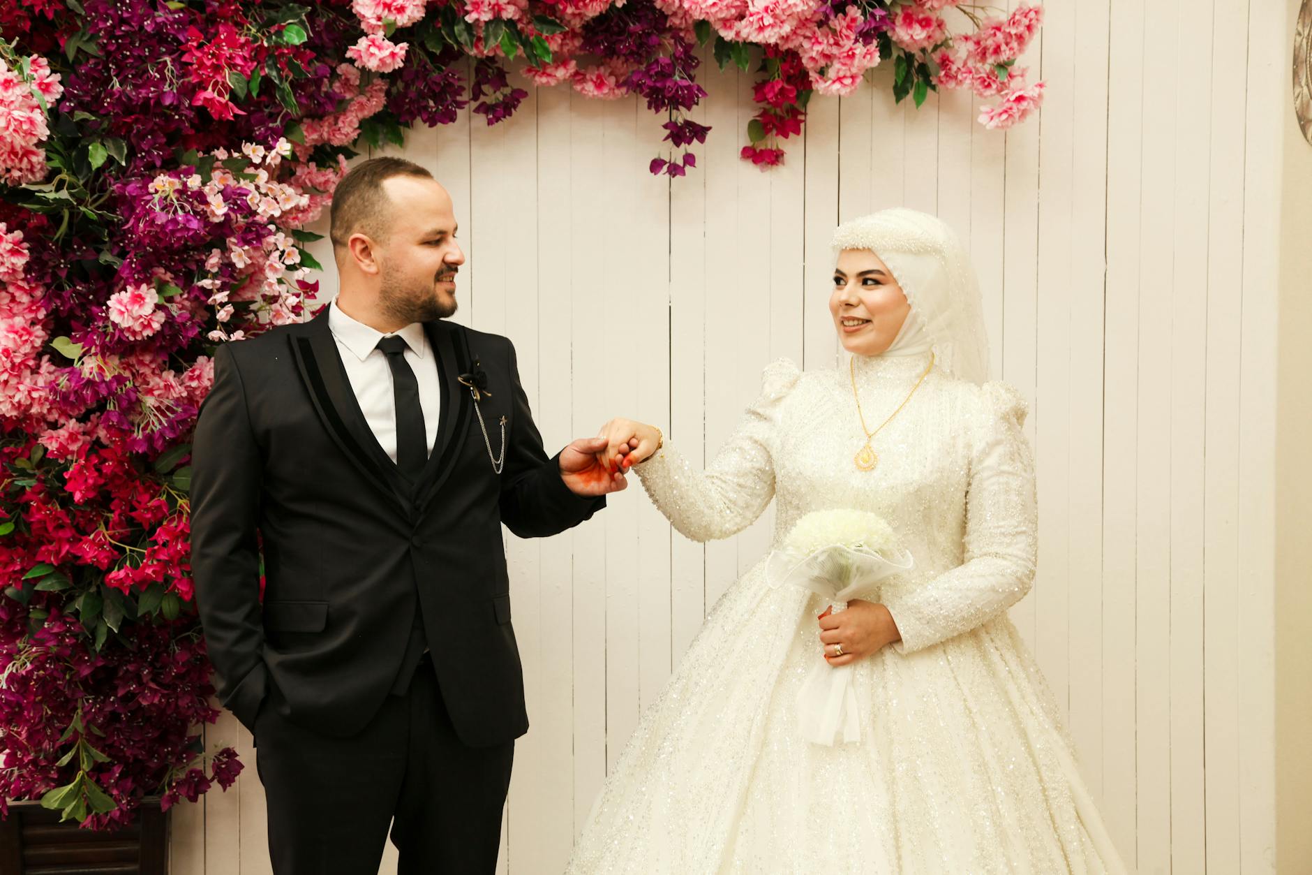 Bride and groom in elegant attire posed with vibrant floral wall backdrop, indoors. - marriage advice bride