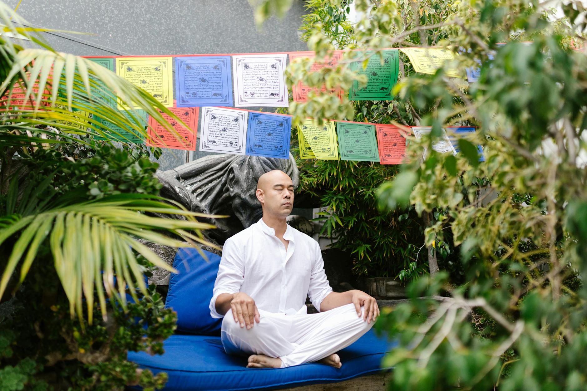 A man in white meditates outdoors in a garden, surrounded by vibrant prayer flags and greenery. - mental clarity techniques