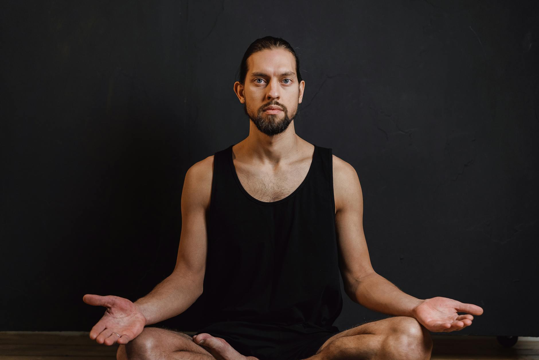 Bearded man meditating in a black tank top against a dark background, promoting peace and wellness. - mindfulness for stress