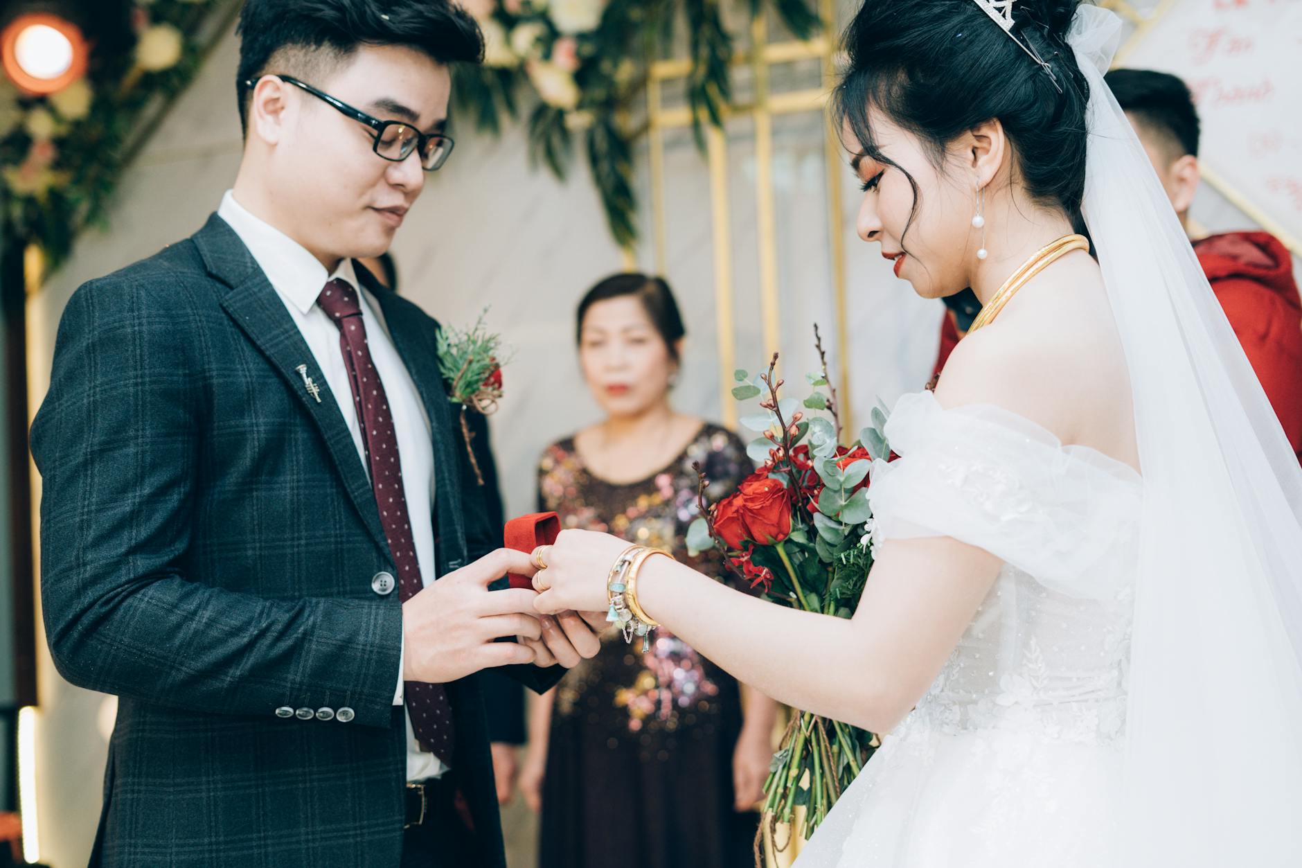 Side view of crop ethnic bride with makeup in dress with bridal bouquet and groom in suit with eyeglasses preparing rings during marriage ceremony in decorated hall behind parent - newlywed marriage advice