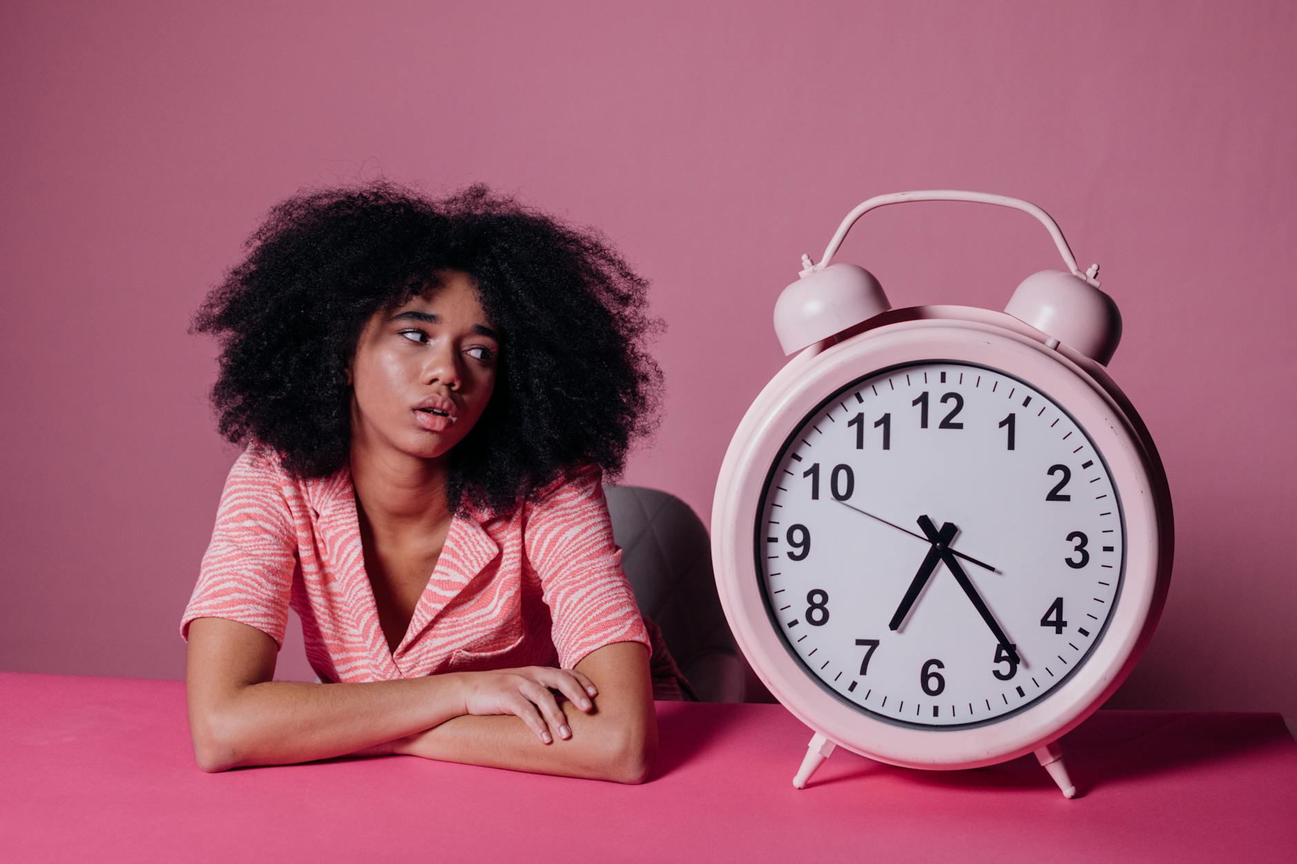 Young woman leaning on table with pink alarm clock, looking pensive. - overcome procrastination