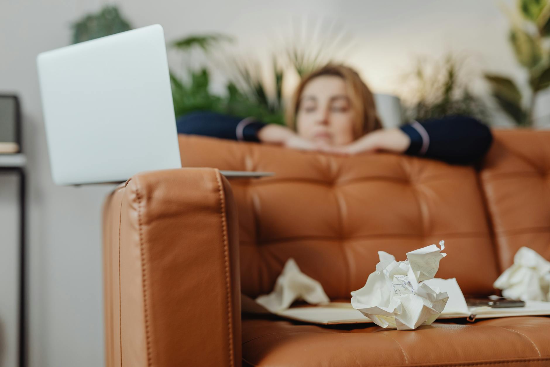 Woman resting on a leather sofa with a laptop and crumpled papers, surrounded by plants. Indoors setting. - overcome procrastination