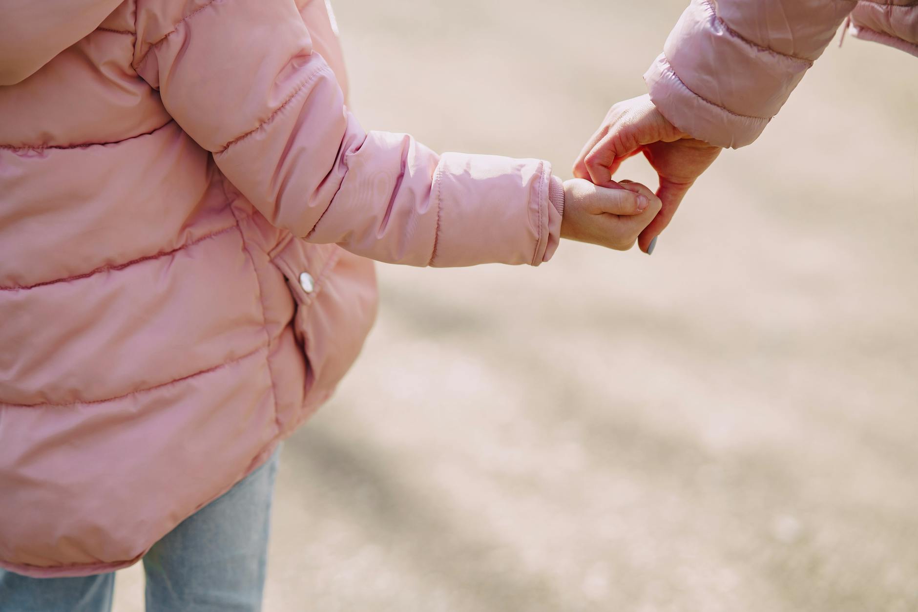 Back view of unrecognizable little girl wearing warm pink jacket holding hands with crop young woman while spending time together in city outside illustrating mother care and support concept - post divorce co parenting