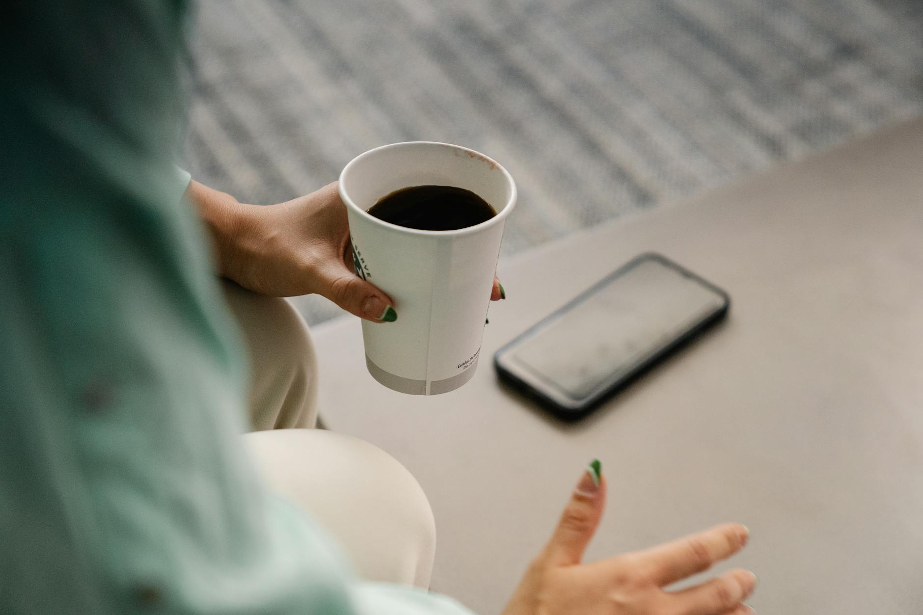Woman holding a paper coffee cup with smartphone nearby in a relaxed setting. - post winter break routine
