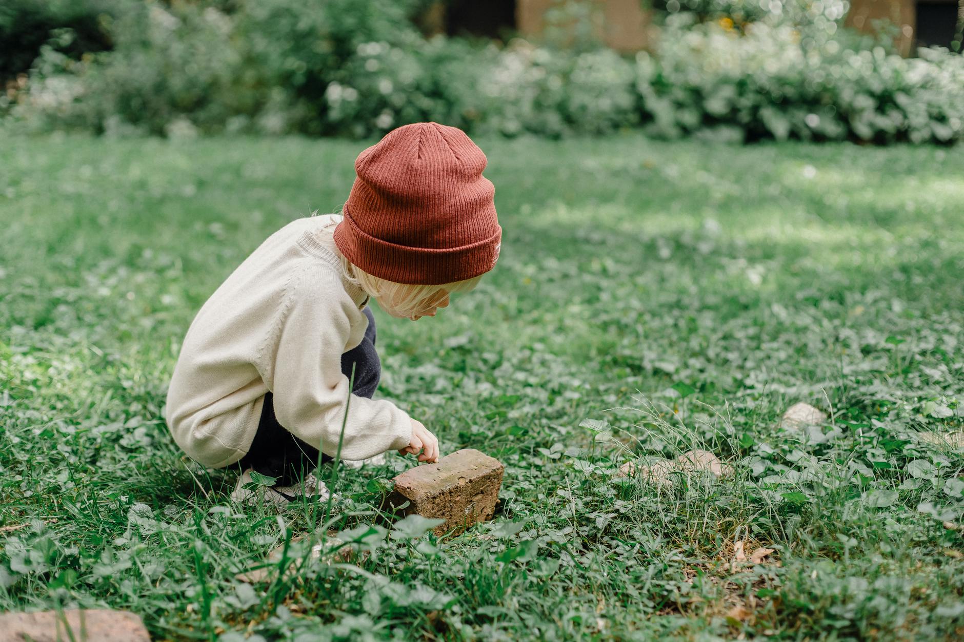 Full body side view of playful kid wearing hat squatting on grassy lawn while exploring small stone against blurred background - preschool nature activities
