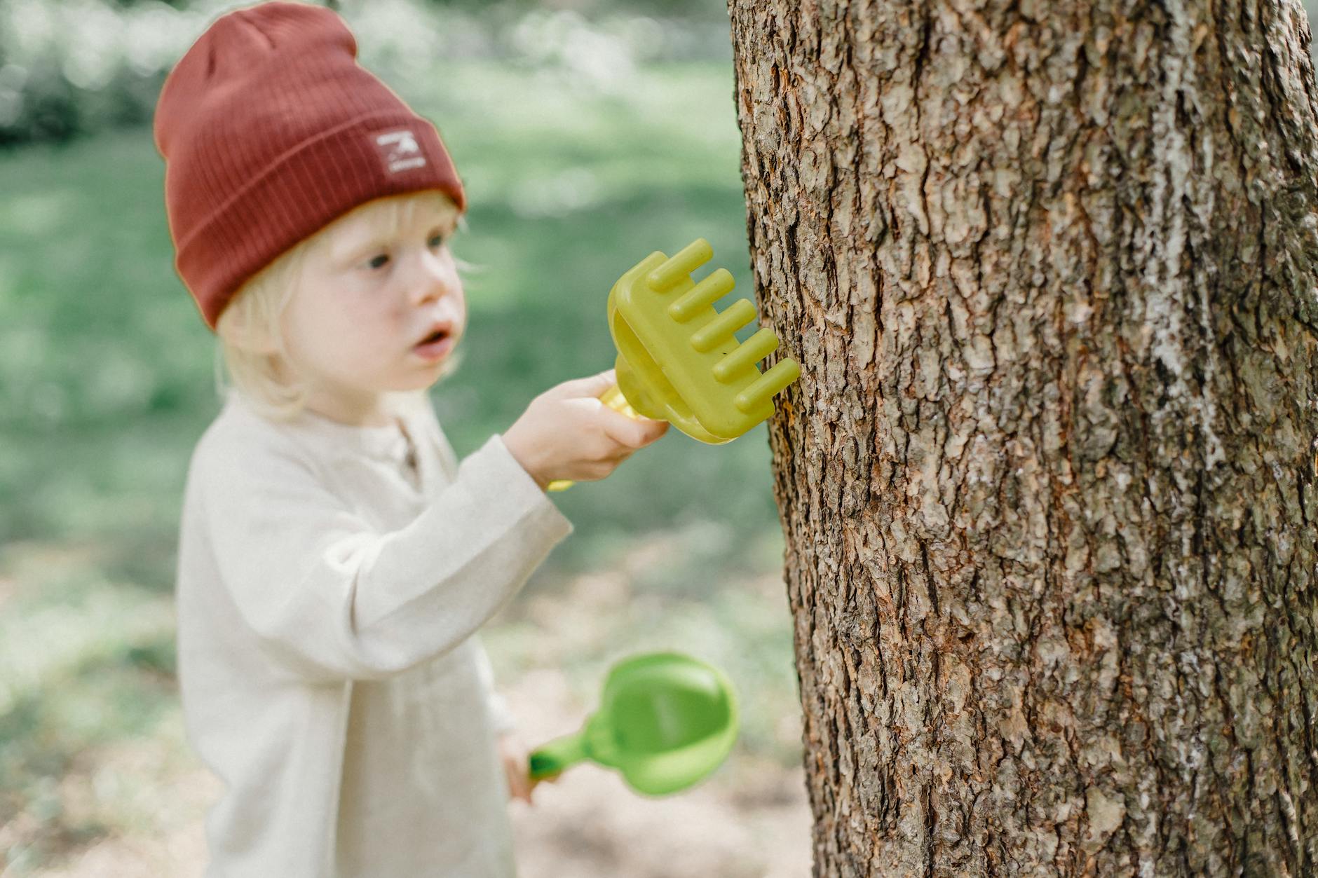 High angle of focused little blond boy in casual wear and brown hat rubbing tree trunk with toy rake in garden - preschool nature activities