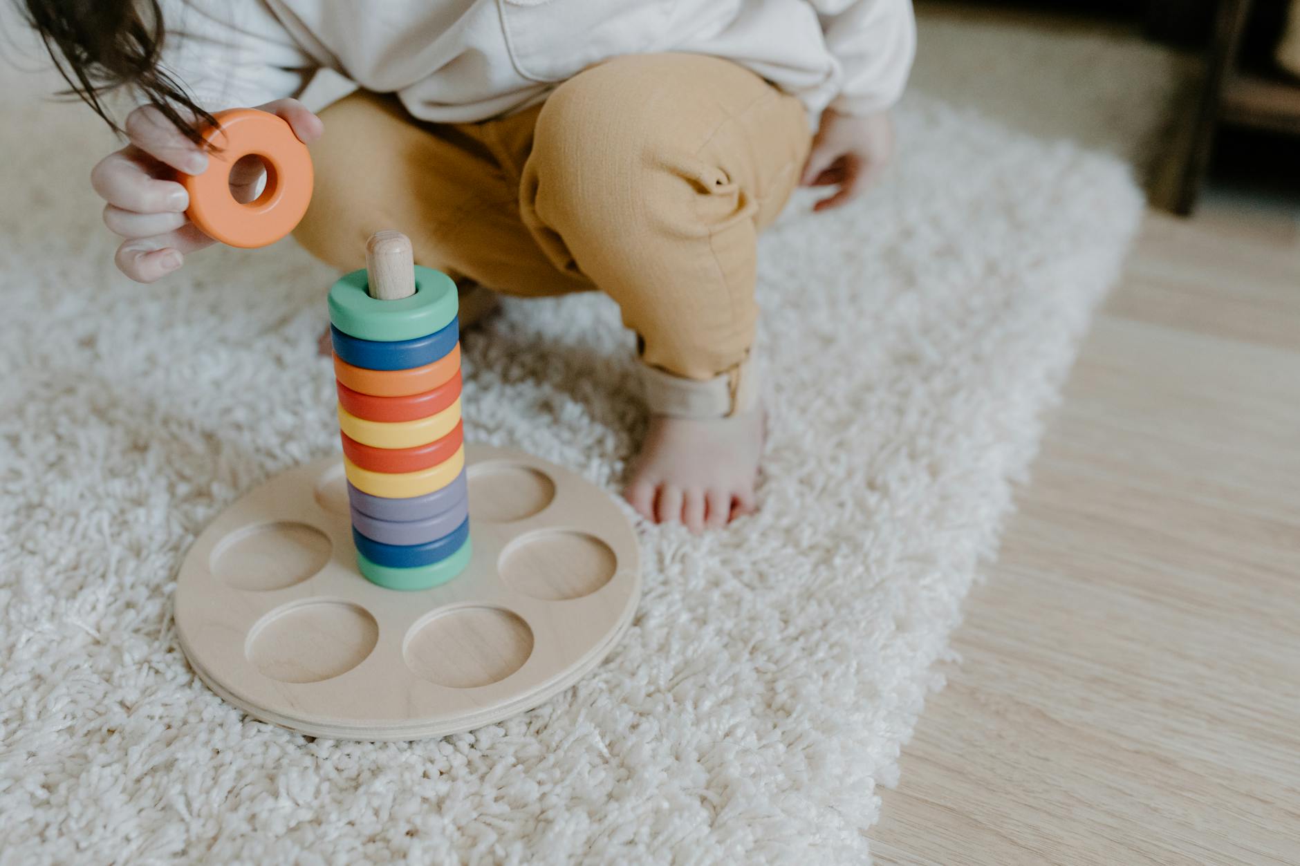 A child playing with a wooden ring stacking toy on a carpet indoors. - preschooler independence