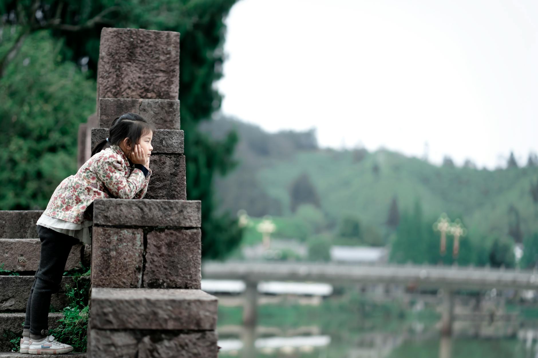 A young girl leans thoughtfully on a stone structure overlooking a serene lakeside view, capturing a moment of contemplation. - preschooler independence