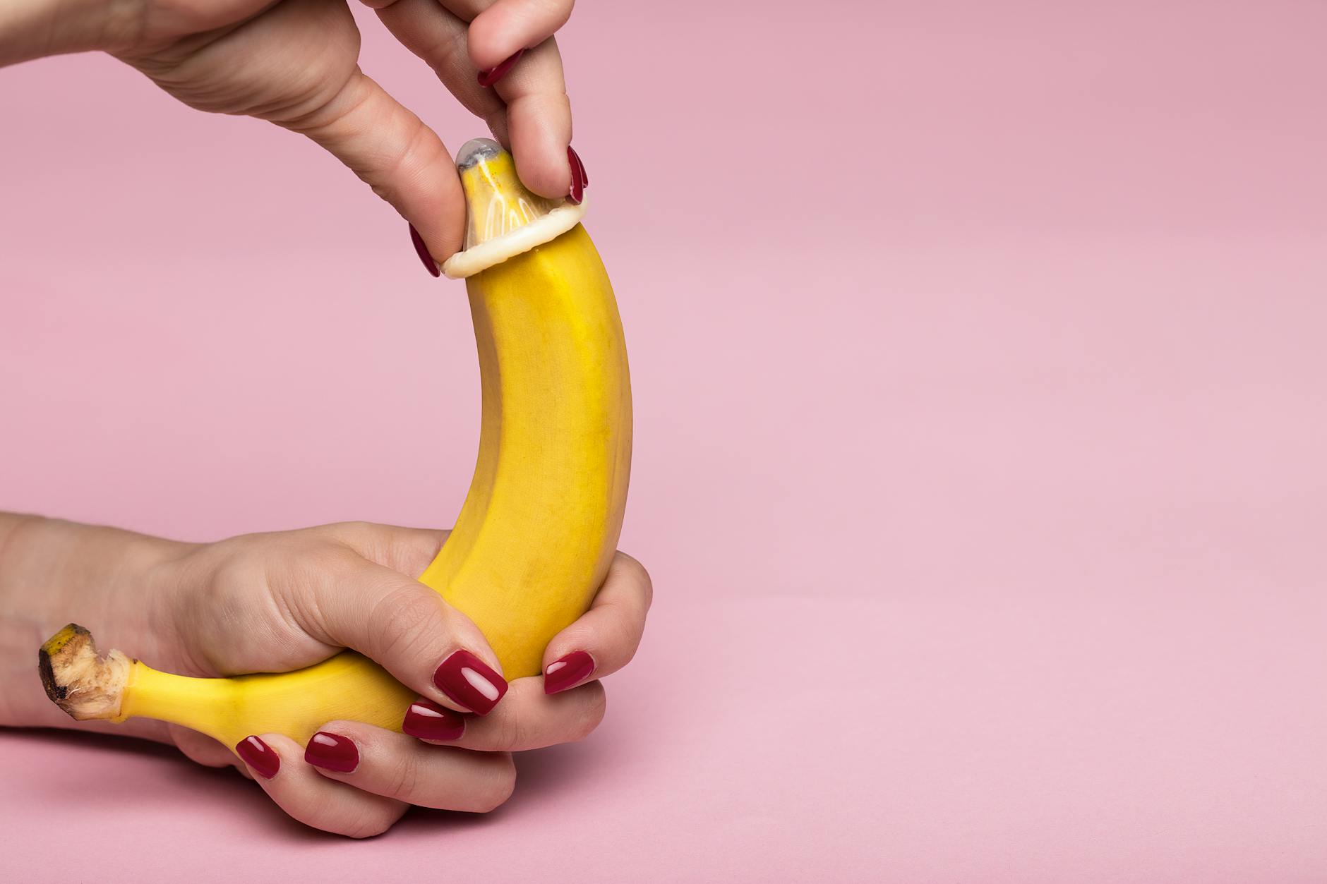 Close-up of hands applying a condom on a banana against a pink background symbolizing safe sex. - prevent spring s.a.d.