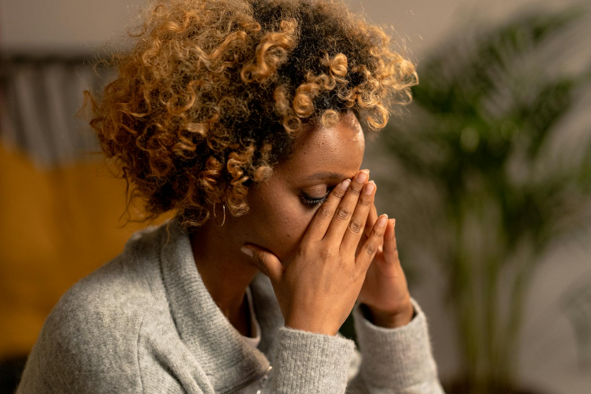 A woman with curly hair in a gray sweater, holding her face, depicting stress indoors. - relationship anxiety reddit
