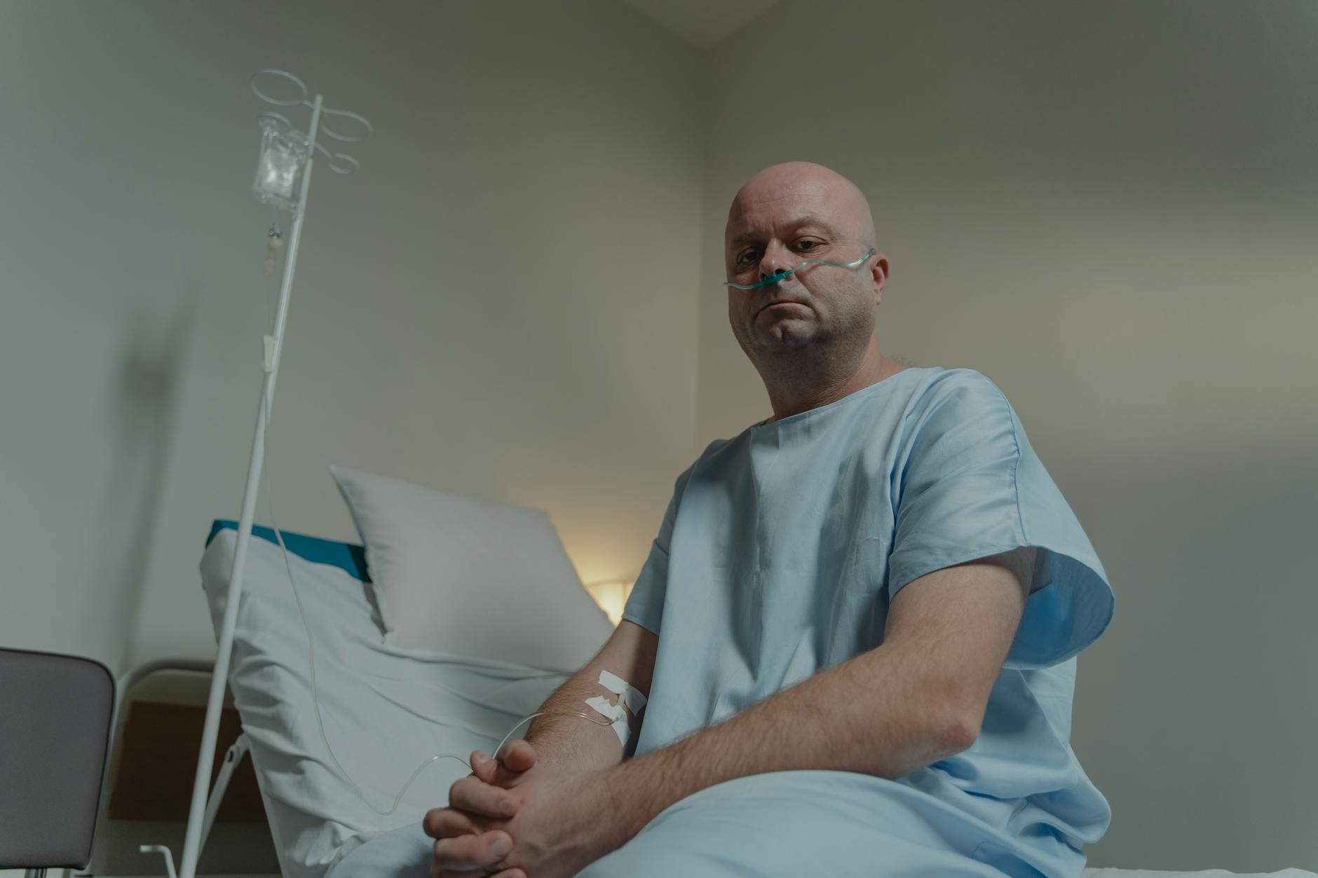 Bald man in hospital gown sitting on bed with nasal cannula and IV in a clinical setting. - sad cbt treatment