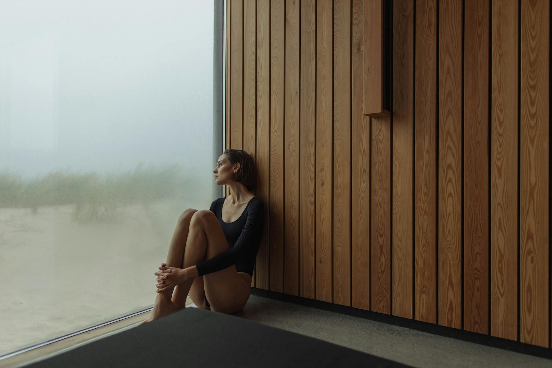 Woman in black longsleeve bodysuit sits contemplatively by a foggy window. - sad spring mood